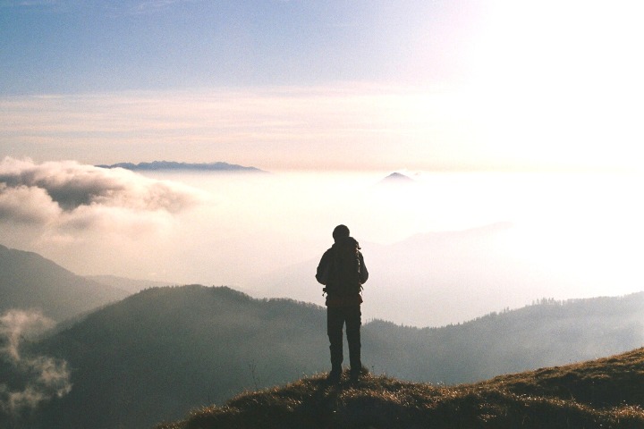 Hiker stands on a mountain, looking into a valley filled with hills and clouds